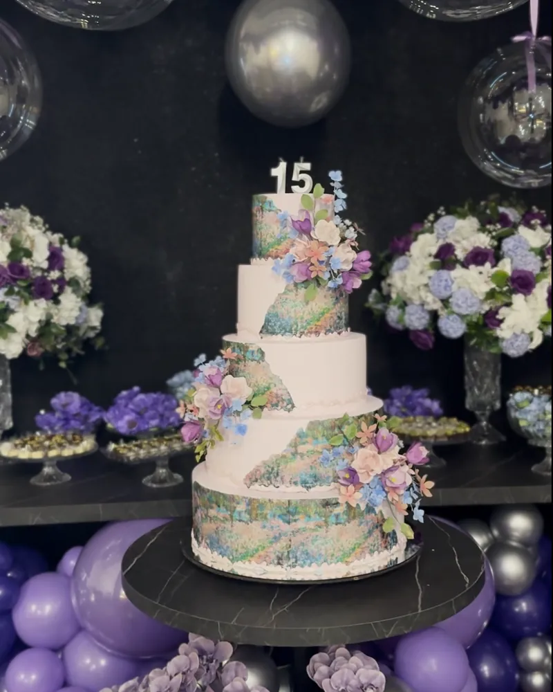 Bolo de 15 anos de três andares com flores lilás, cercado de balões e arranjos na festa de debutante