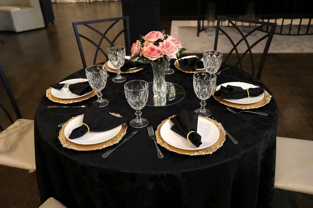 Mesa redonda posta com toalha preta, sousplats dourados, taças de cristal e arranjo de rosas na Ônix Eventos
