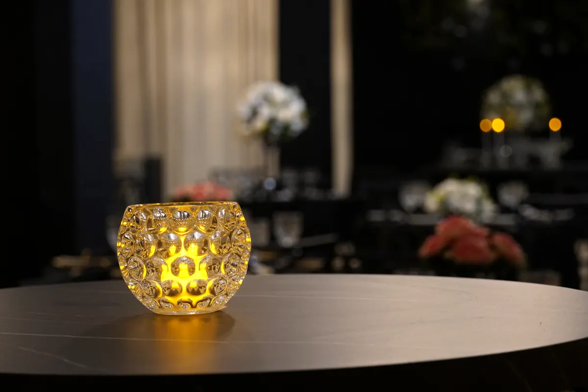 Luminária de vidro com luz quente sobre mesa, com o salão decorado ao fundo na Ônix Eventos